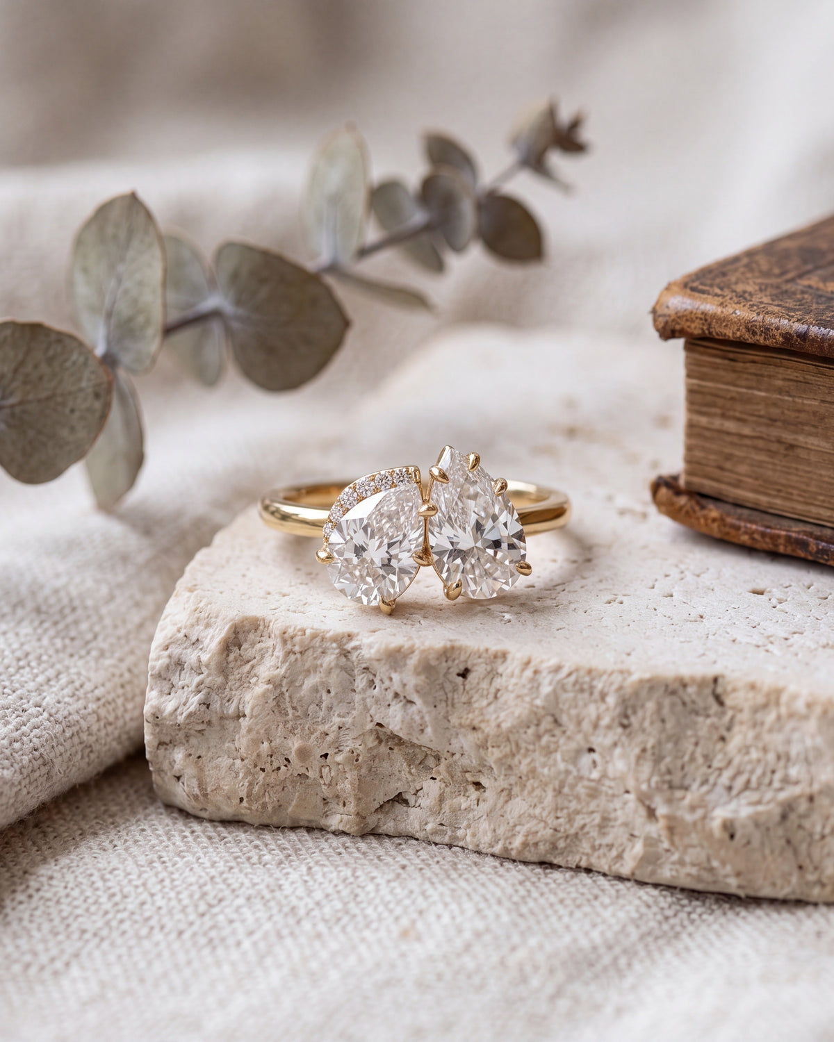 A gold double-stone ring with round brilliant-cut clear gemstones rests on a textured stone slab, styled with eucalyptus leaves and a vintage book on a linen fabric background.