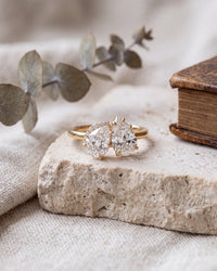 A gold double-stone ring with round brilliant-cut clear gemstones rests on a textured stone slab, styled with eucalyptus leaves and a vintage book on a linen fabric background.