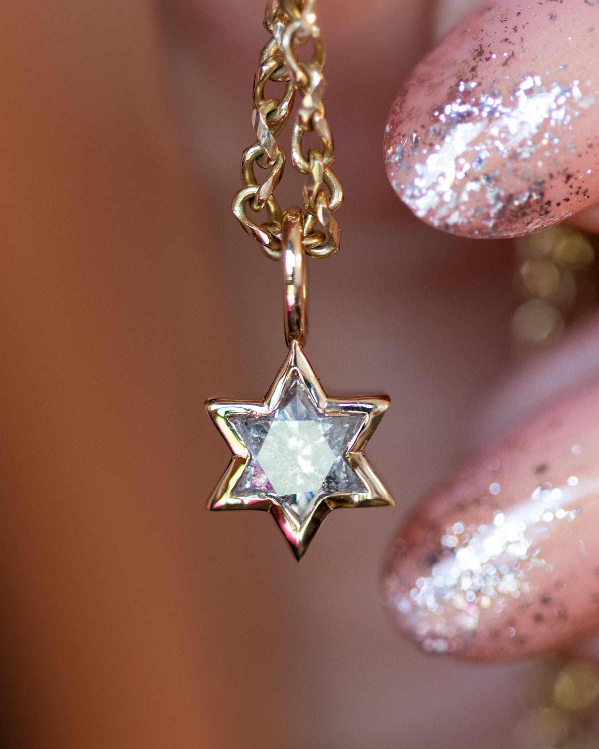 A close-up of a gold Star of David pendant necklace with a clear faceted gemstone, held by fingers with glittery pink nails against a warm, blurred background.