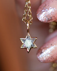 A close-up of a gold Star of David pendant necklace with a clear faceted gemstone, held by fingers with glittery pink nails against a warm, blurred background.