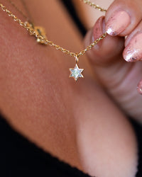 A close-up lifestyle shot of a delicate gold chain necklace with a small star-shaped clear gemstone pendant, worn on a person's neck and held by a hand with glittery pink nails.